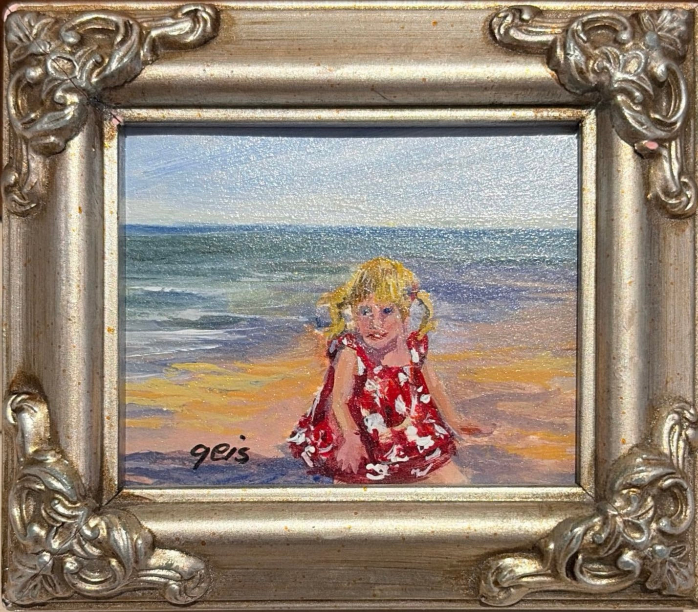 Girl on Beach by Kay Geis, original miniature oil painting of a young blonde girl in a red floral dress sitting on a sandy beach beside the ocean in an ornate silver frame.